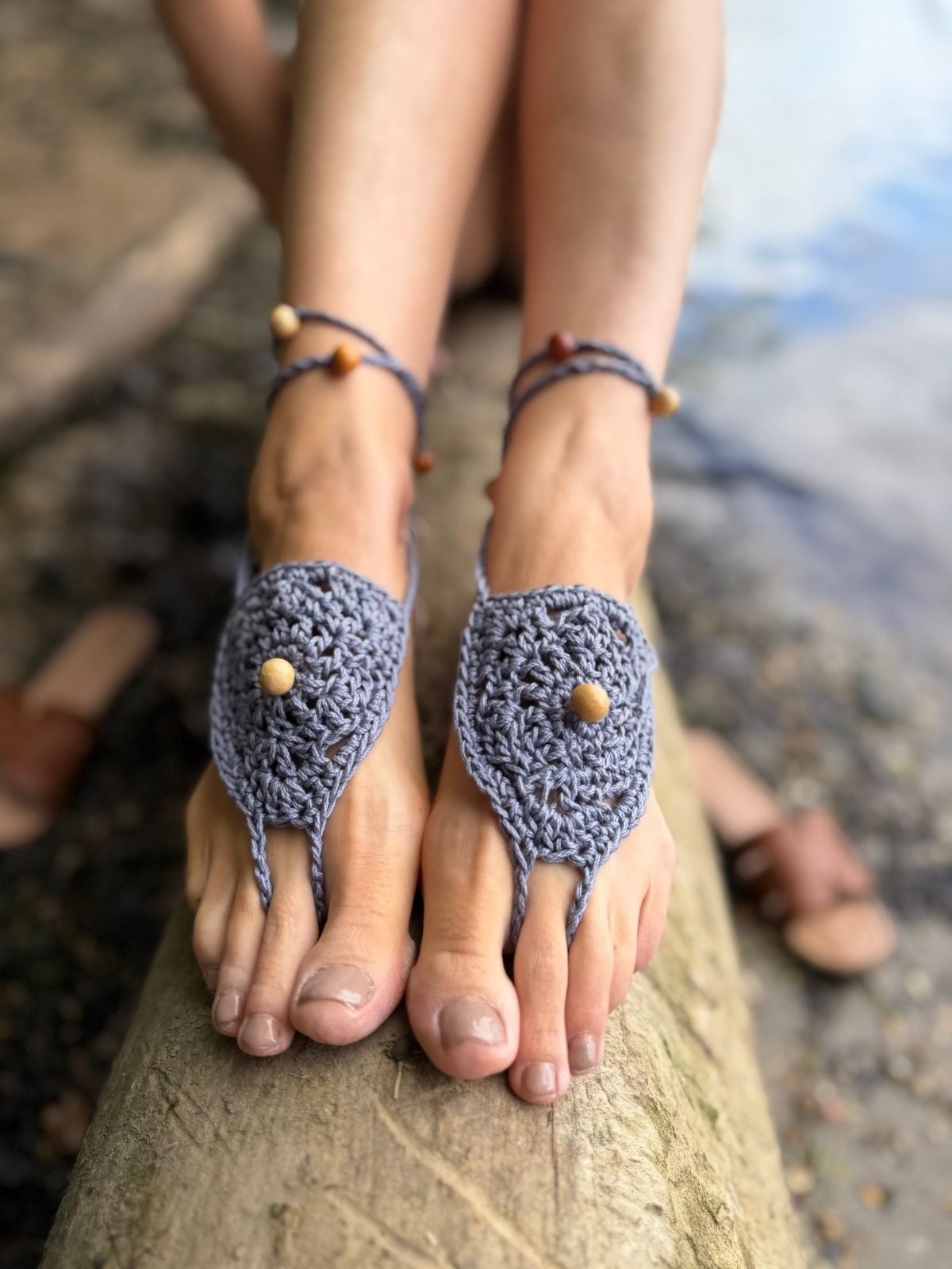 Person wearing handmade crochet barefoot sandals with wooden beads, sitting on a log near water. Toes and feet are visible, with another pair of sandals in the background—created using a free barefoot crochet sandals pattern.