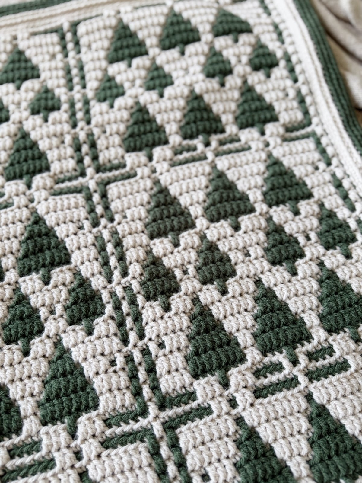 Close-up of a Christmas crochet afghan, showcasing a repeating pattern of green triangles and beige squares with textured stitches and a striking geometric design.