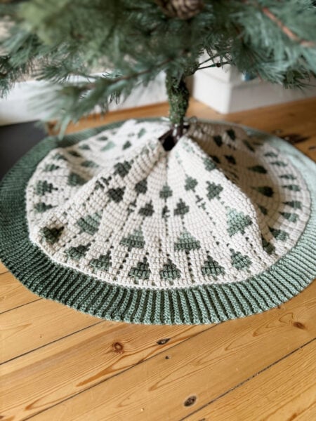 Crocheted tree skirt with green and white geometric pattern and ribbed edge beneath a Christmas tree on a wooden floor.