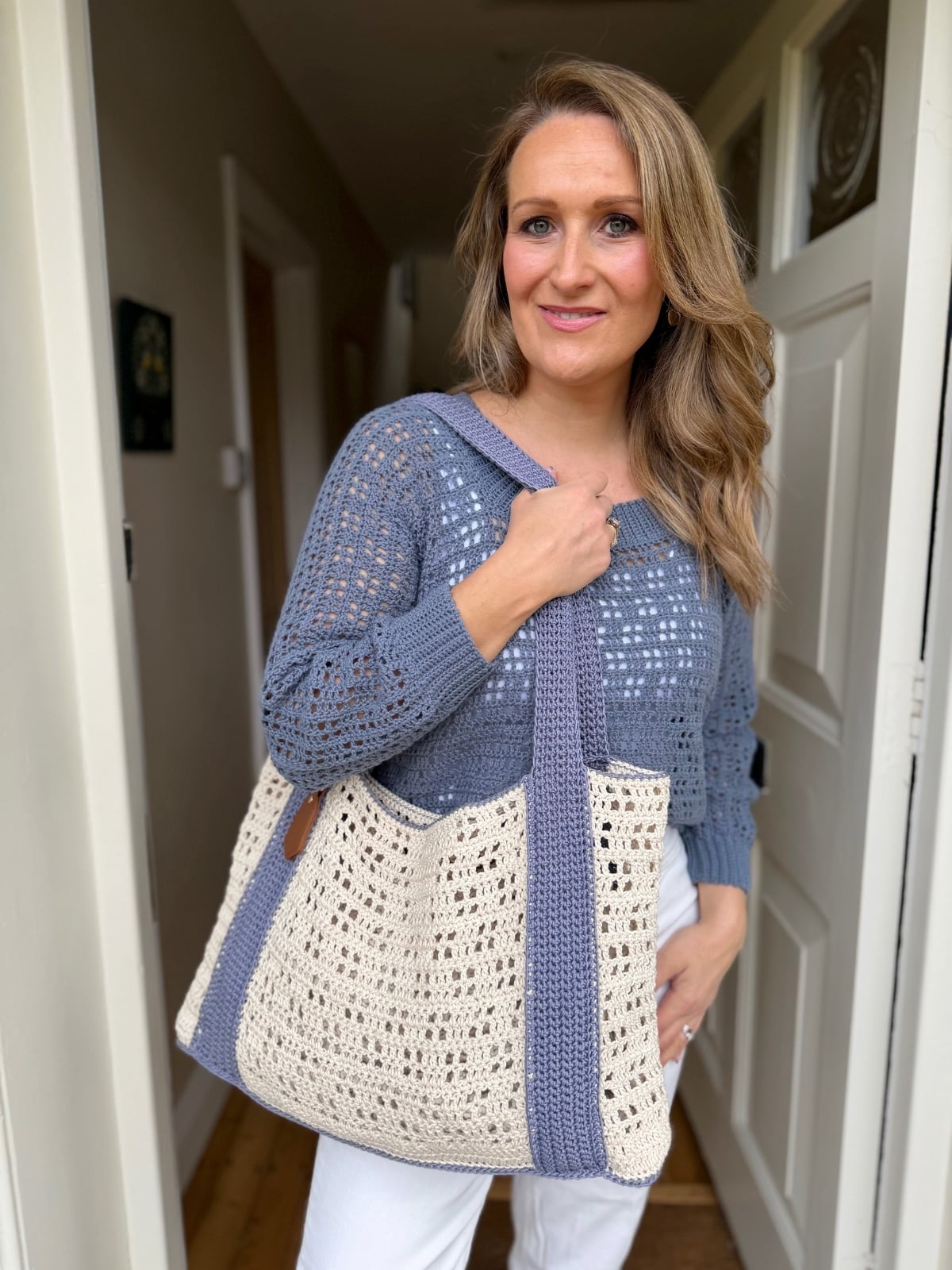 Woman standing in a doorway, wearing a blue knit sweater and white pants, holding a large crochet tote bag with cream and gray colors made from a free crochet beach bag pattern.