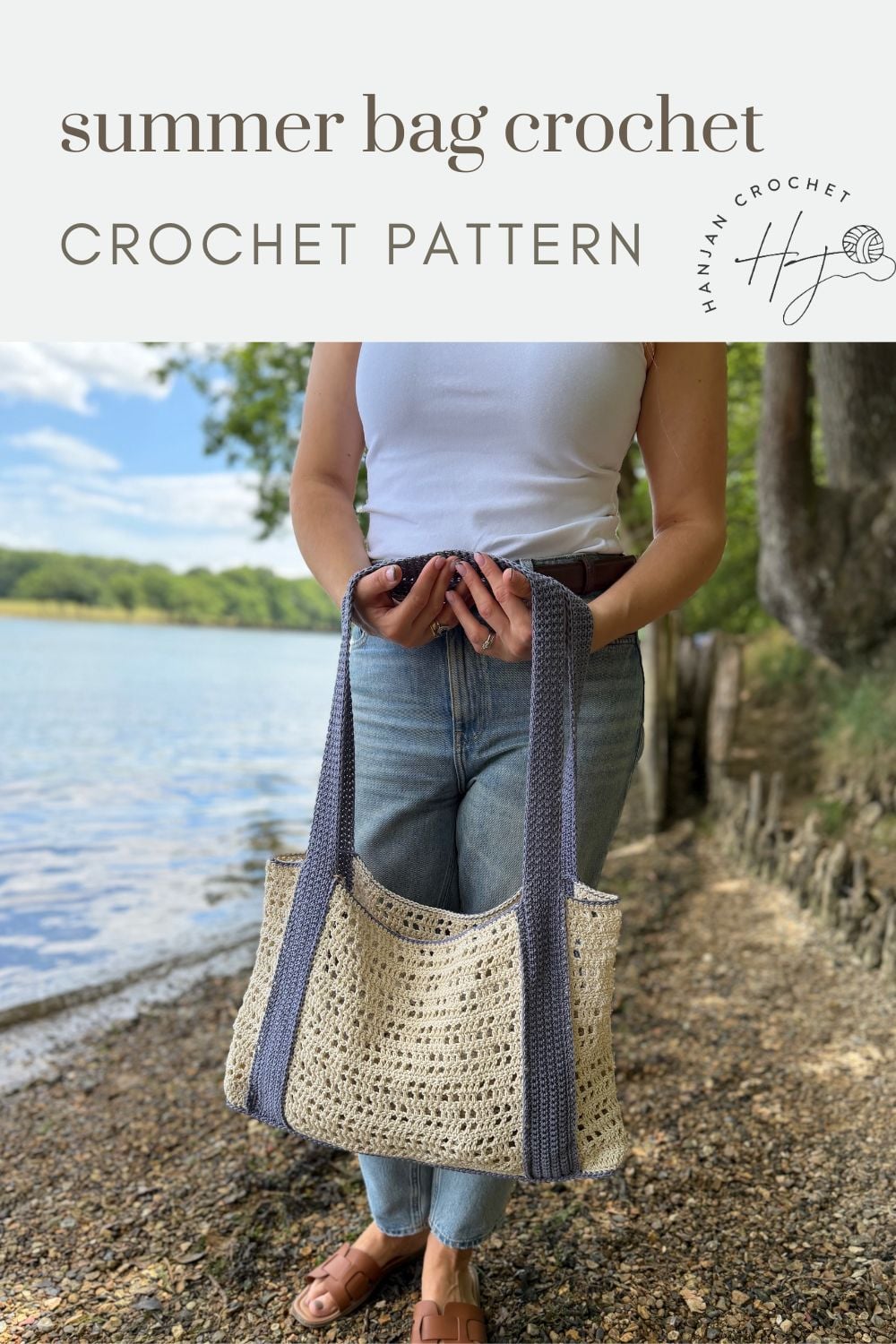 A person stands by a lakeshore holding a handmade crochet bag with dark handles, wearing jeans and a white tank top. Text reads "summer bag crochet CROCHET PATTERN—perfect for those seeking a free crochet beach bag pattern.