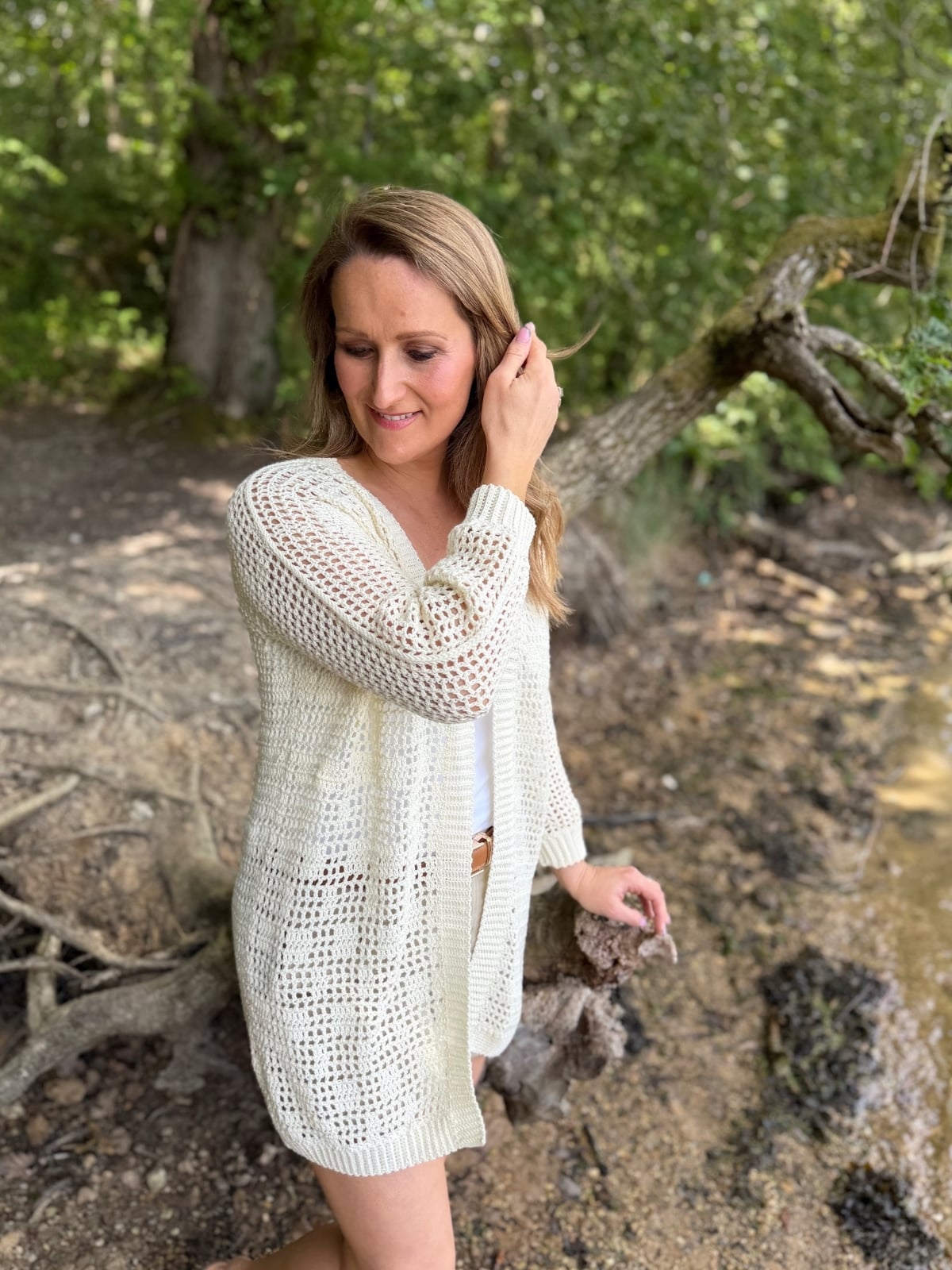 A woman in a white summer cardigan crochet pattern stands by a wooded lakeshore, touching her hair and looking down with trees and branches in the background.
