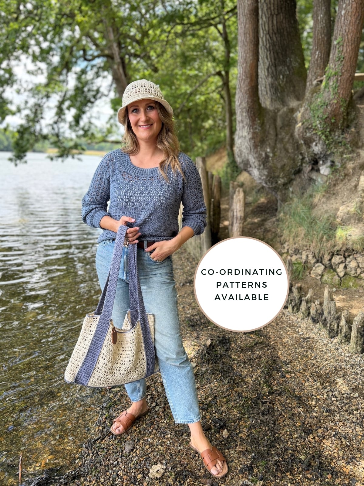 A woman stands by a lake wearing a summer sweater crochet pattern hat, blue sweater, jeans, sandals, and holding a crochet tote bag. Text overlay reads, "Co-ordinating patterns available.