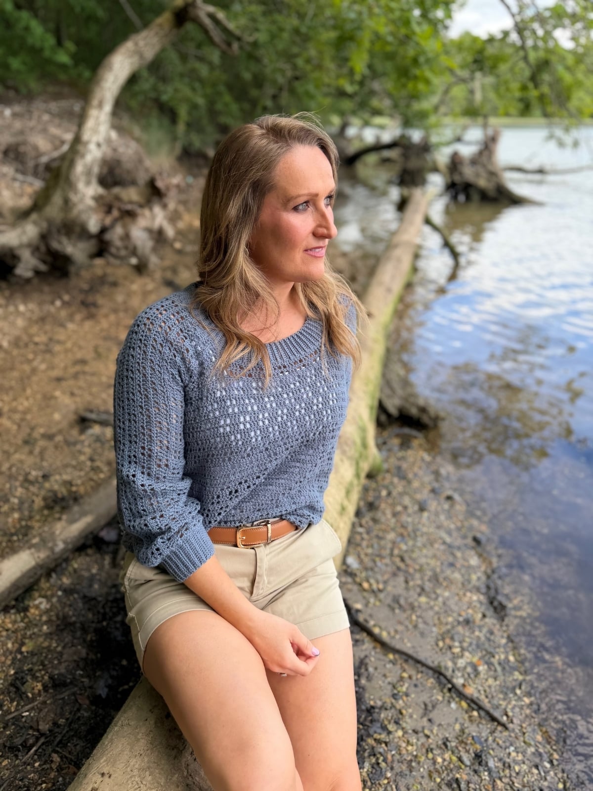 A woman in a blue summer sweater crochet pattern and beige shorts sits on a large log by a lakeshore, looking into the distance with trees and water in the background.