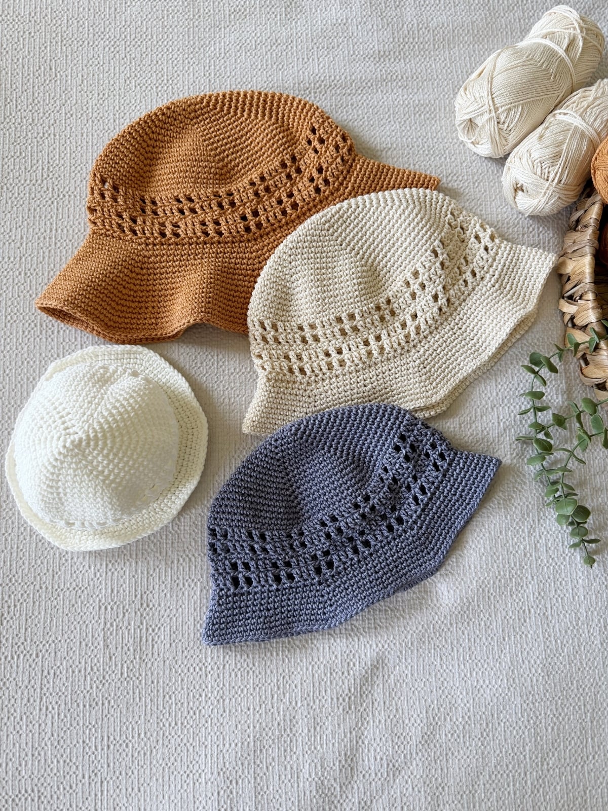 Four crocheted hats in brown, cream, white, and gray are displayed on a textured surface near several balls of yarn and a sprig of greenery—perfect inspiration if you’re searching for a free crochet sun hat pattern.