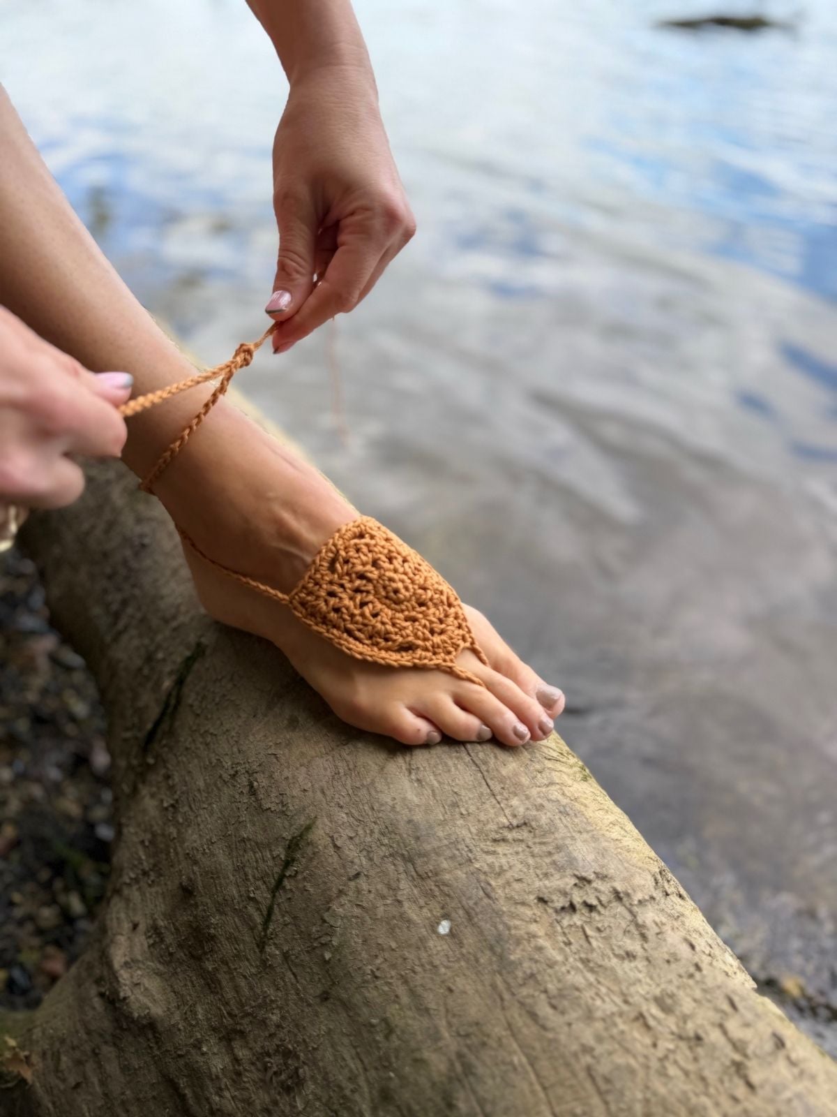 A person ties a brown crocheted barefoot sandal onto their foot while sitting on a large log near the water's edge, using a free barefoot crochet sandals pattern for inspiration.