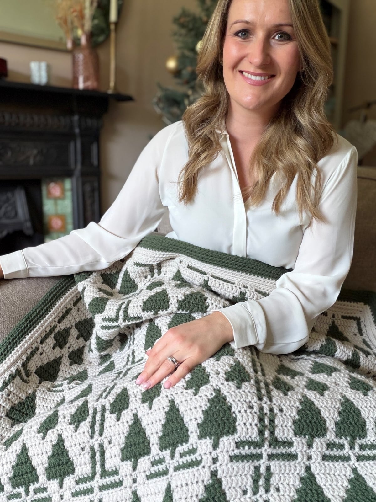 A woman in a white blouse sits on a couch with a Christmas crochet afghan featuring green and white geometric patterns on her lap, smiling at the camera.