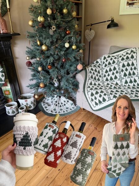 A living room with a decorated Christmas tree, crochet tree-themed decorations, bottle bags, a cup cozy, and a woman holding a crocheted wine bottle bag.