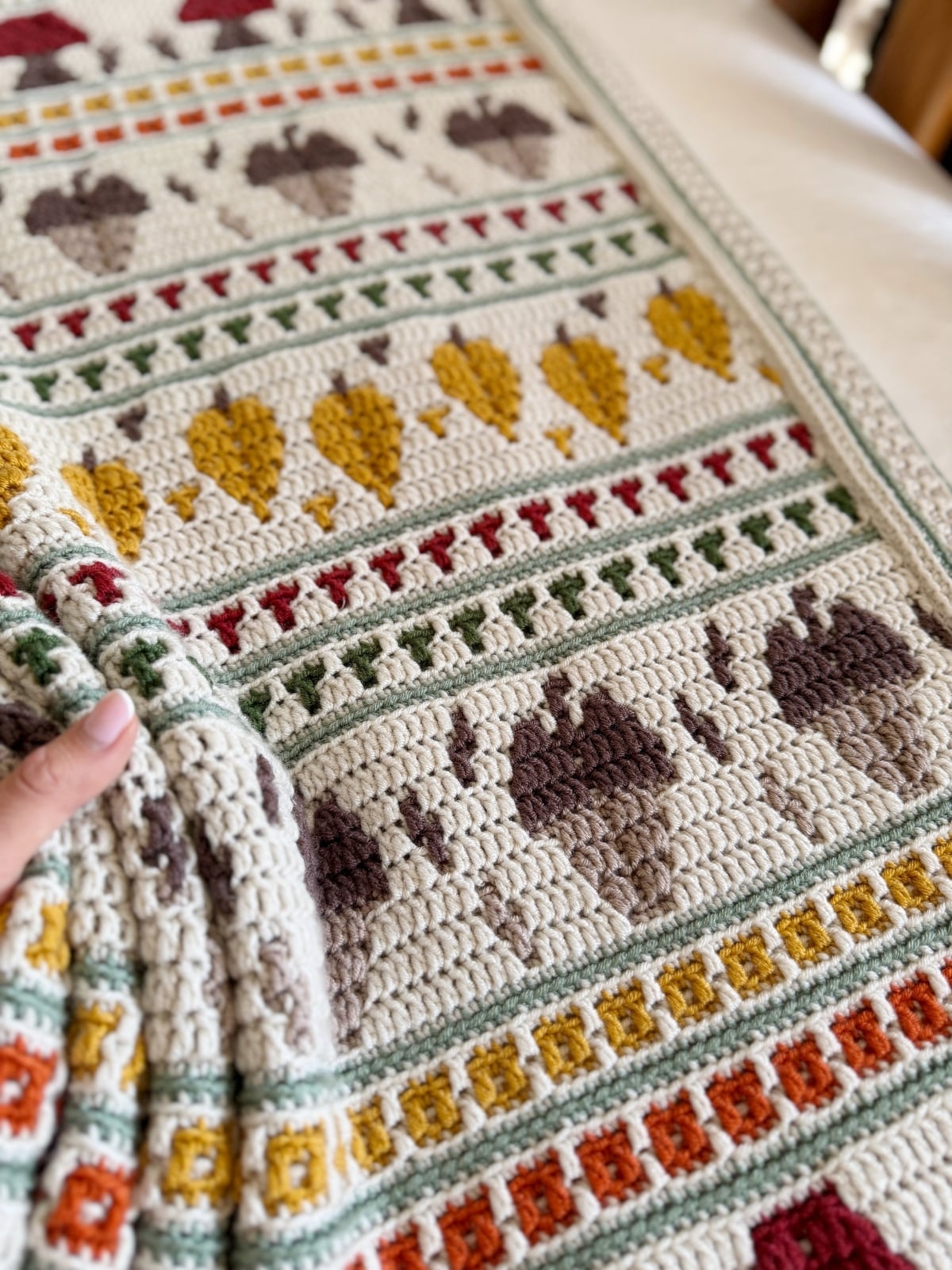A hand holds a crocheted blanket featuring rows of geometric and nature-inspired patterns in earthy tones, including leaves and bear paw motifs.