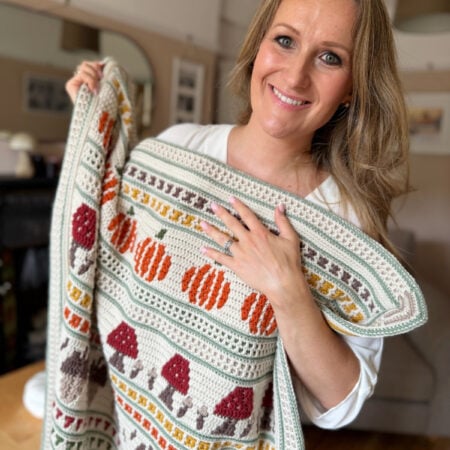 Woman smiling and holding a colorful knitted blanket with patterns of pumpkins, mushrooms, and leaves in a cozy living room.