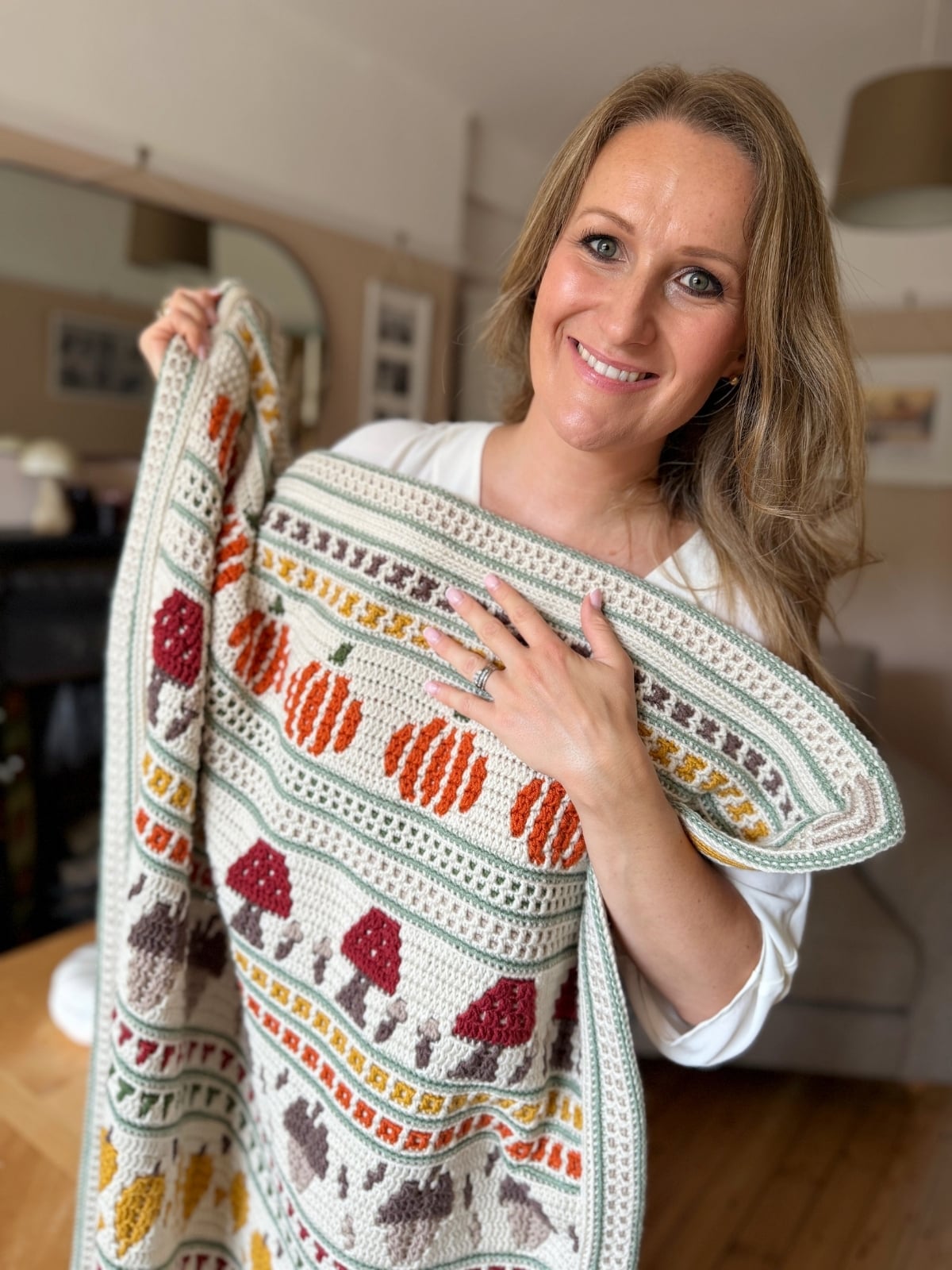 Woman smiling and holding a colorful knitted blanket with patterns of pumpkins, mushrooms, and leaves in a cozy living room.