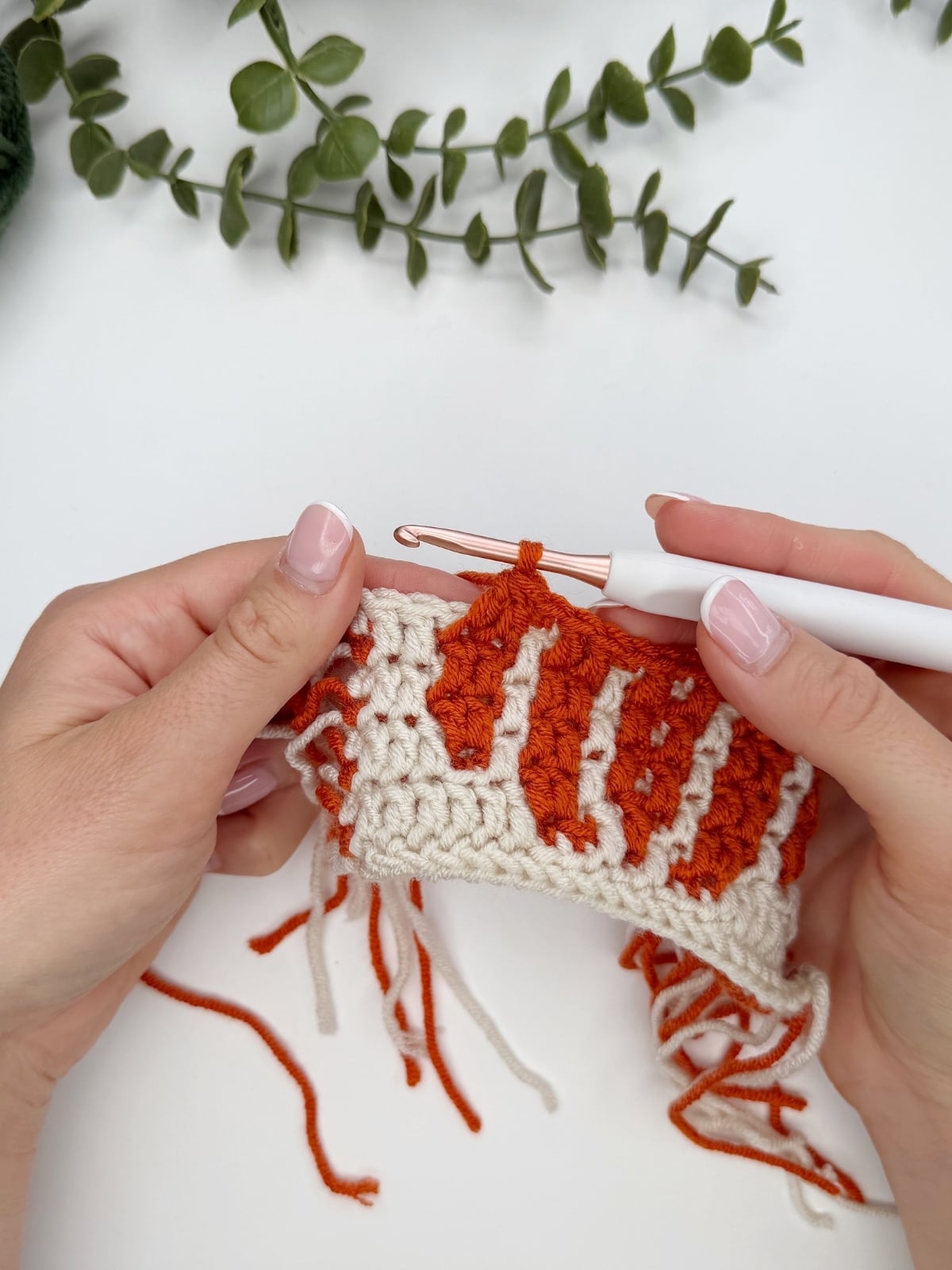 Two hands crochet a white and orange patterned piece with a white crochet hook. Loose yarn ends are visible, and a green plant is in the background.