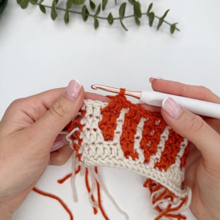 Hands holding a crochet project with orange and white yarn, using a white crochet hook, with green leaves in the background on a white surface.