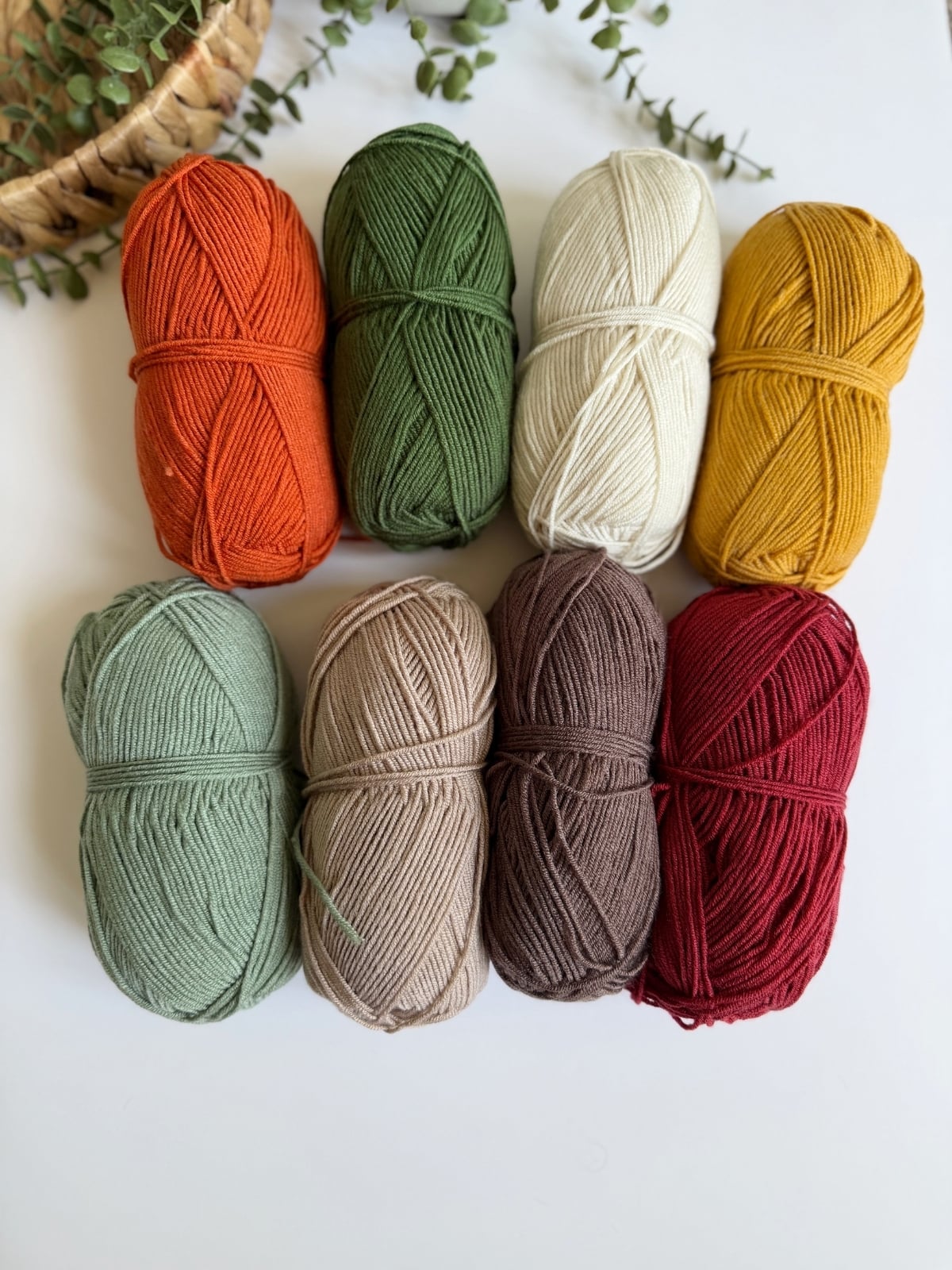 Eight skeins of yarn in various colors, including orange, green, cream, yellow, mint, tan, brown, and red, arranged neatly in two rows on a white surface.
