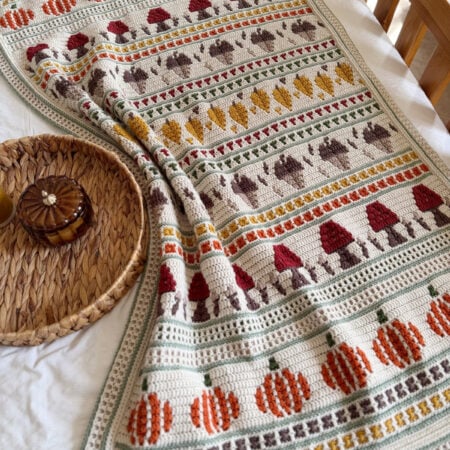 Crocheted blanket with rows of pumpkins, mushrooms, and leaves in orange, red, yellow, and brown on a cream background, draped over a bed near a round woven tray.