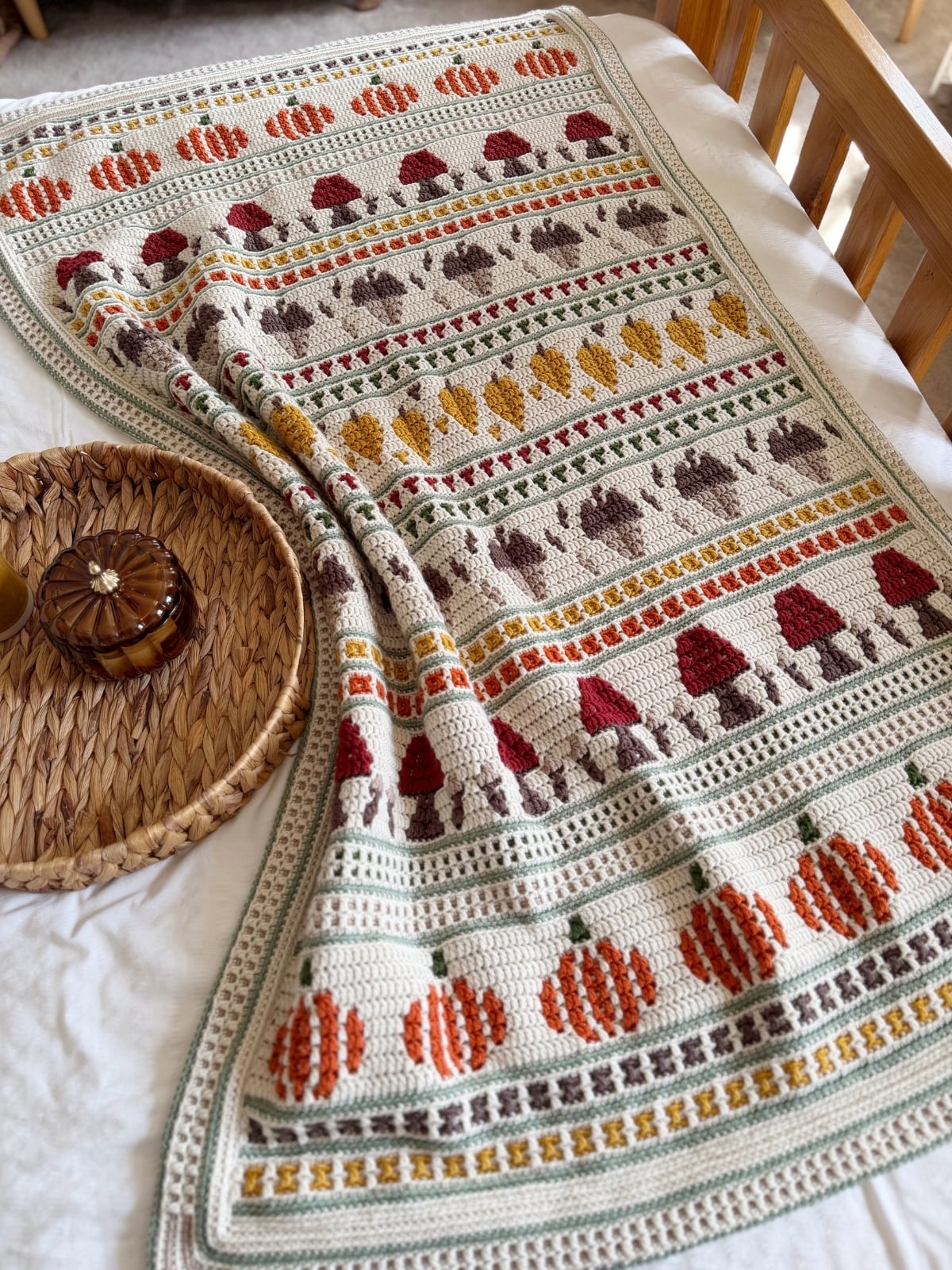 Crocheted blanket with rows of pumpkins, mushrooms, and leaves in orange, red, yellow, and brown on a cream background, draped over a bed near a round woven tray.