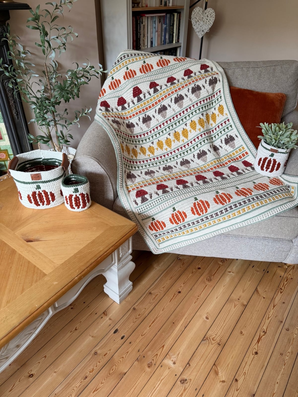 A beige armchair displays a crocheted blanket with fall motifs along with matching crochet baskets and a potted plant on a wooden table nearby.