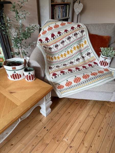 A crocheted blanket with autumn leaf and pumpkin patterns drapes over a beige armchair, next to matching crochet baskets on a wooden table and a potted plant.