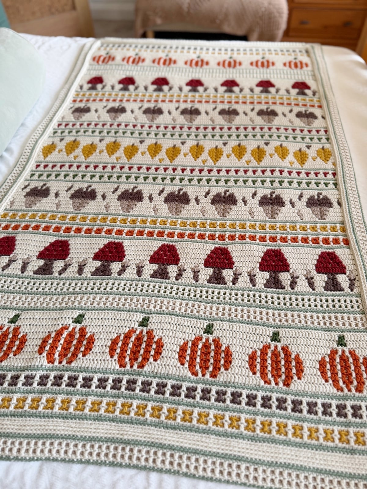 A crocheted blanket with rows of pumpkins, mushrooms, and geometric patterns in orange, brown, yellow, and green on a white background.