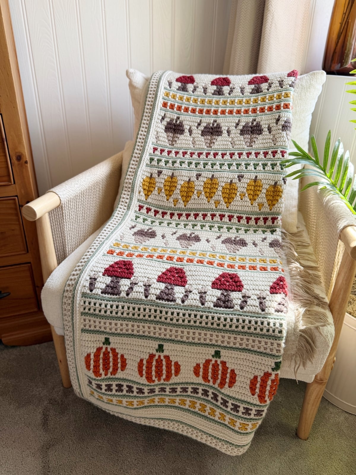 A crocheted blanket with rows of pumpkins, mushrooms, acorns, and leaves in autumn colors draped over a cream armchair in a cozy room.