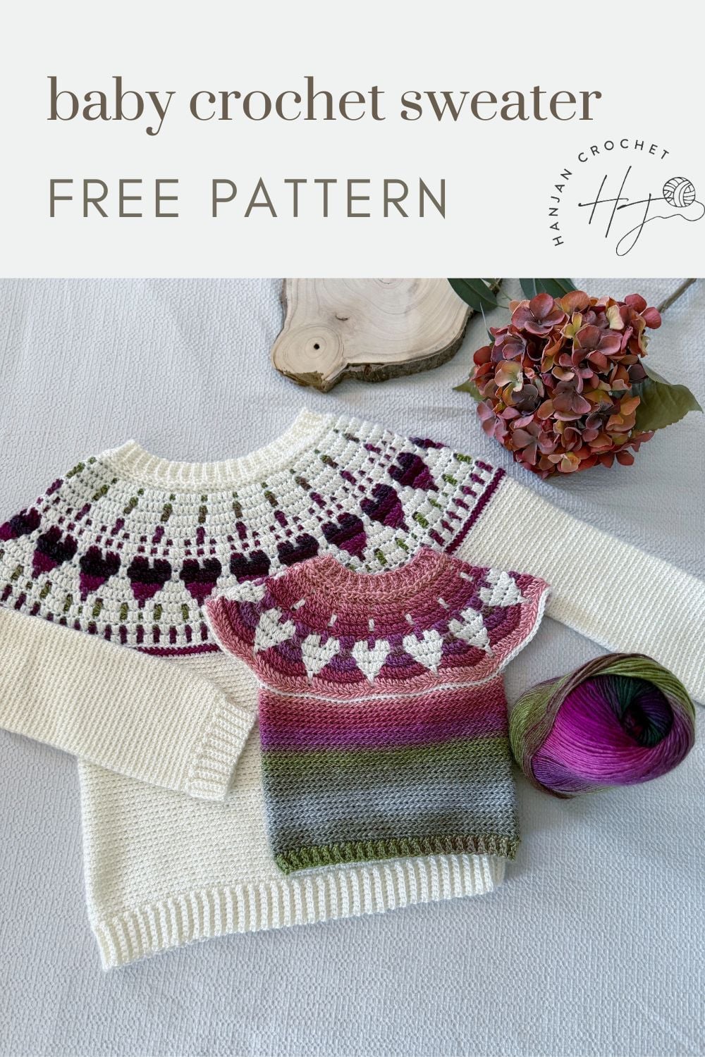 Two crocheted baby sweaters with a heart pattern are displayed on a table with a ball of yarn, a wooden tray, and dried flowers. Text above reads, “baby crochet sweater FREE PATTERN.”.