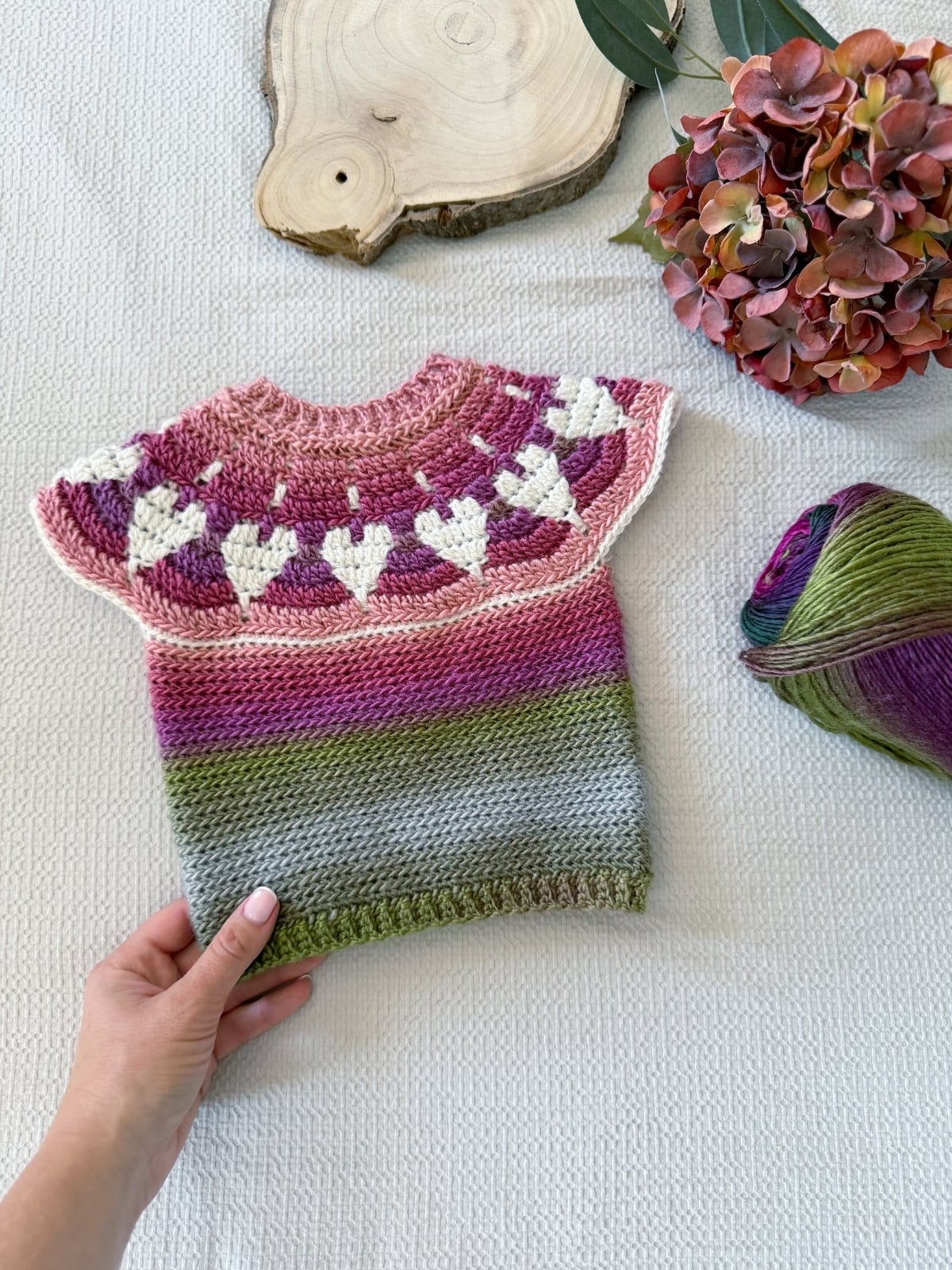 A hand holds a charming baby crochet sweater pattern, featuring heart motifs in pink, white, green, and purple. The cozy knitted piece rests beside a ball of yarn, delicate flowers, and a rustic wooden board.