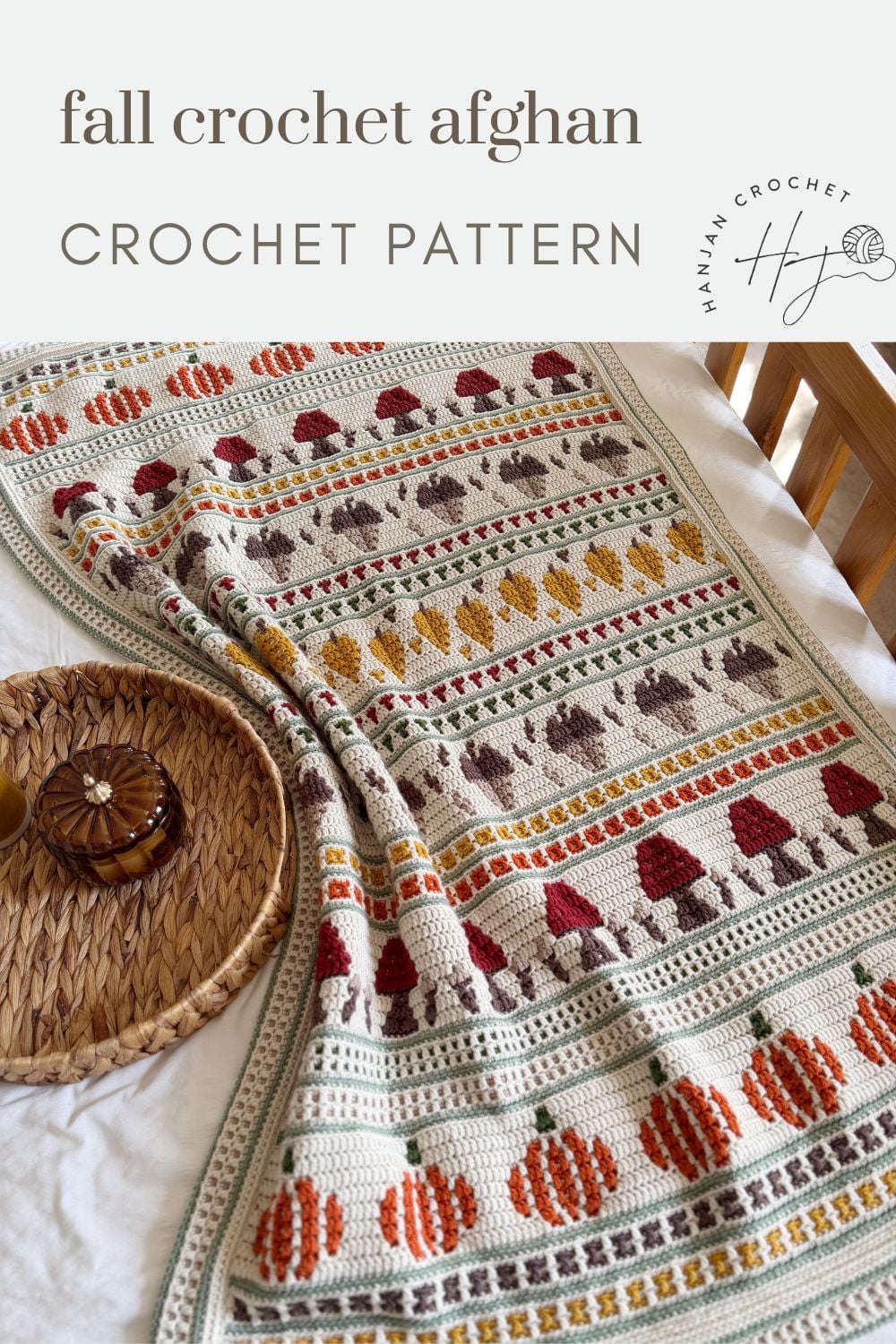 A crocheted afghan with rows of pumpkins, mushrooms, and leaves in fall colors, displayed on a bed next to a woven tray with a drink and coaster.