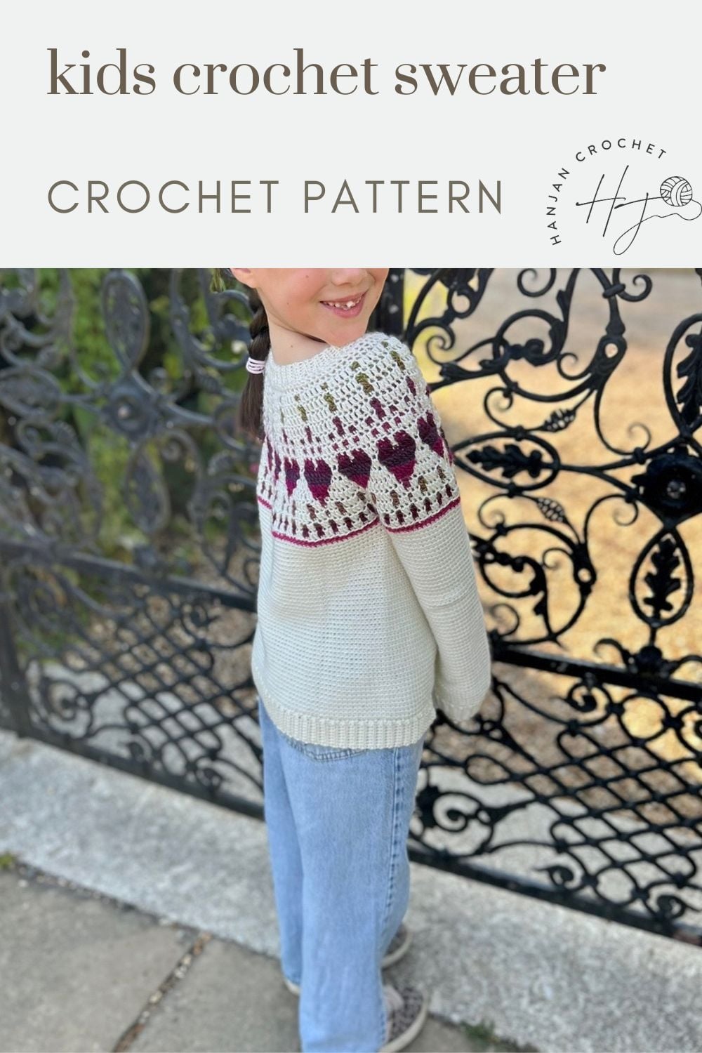 Child wearing a white crochet sweater with maroon heart pattern stands in front of a decorative black gate.