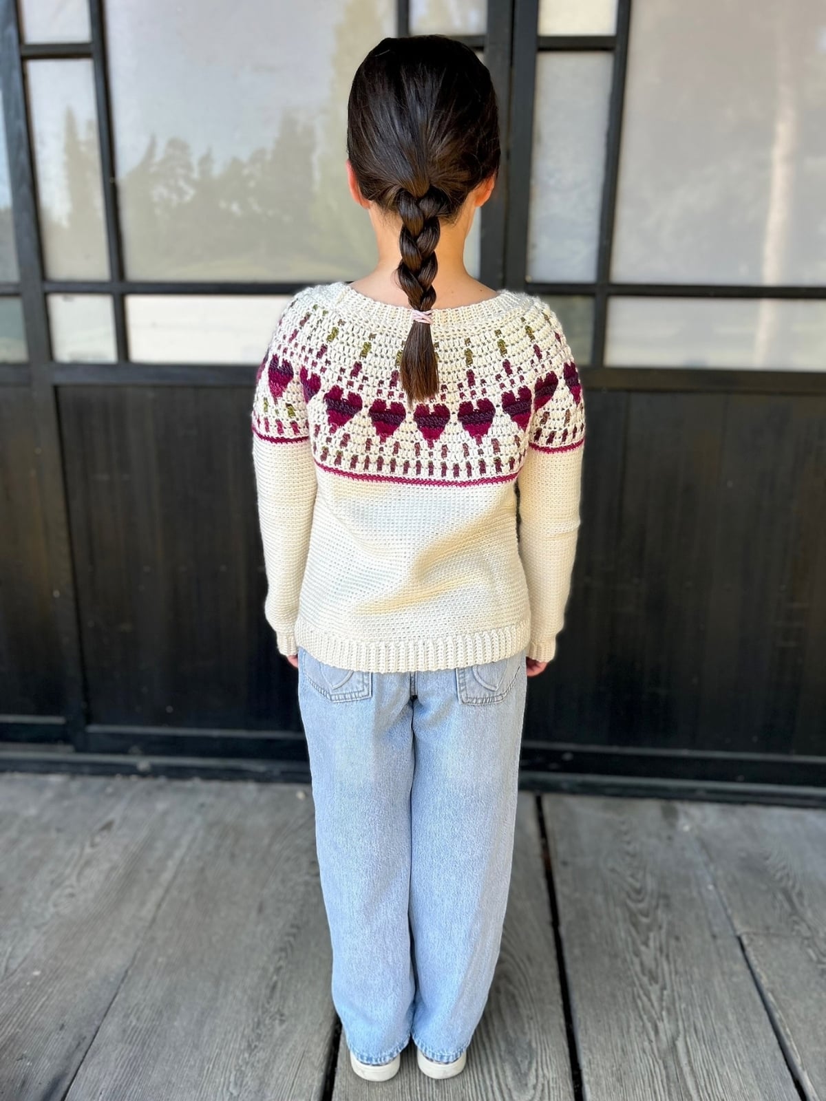 Child with a brown braid is wearing a cream sweater with a red and green pattern inspired by a baby crochet sweater pattern and light blue jeans, standing on a wooden floor facing away from the camera.