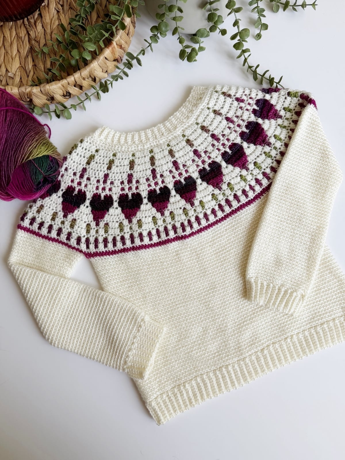 A cream-colored knit sweater with a patterned yoke featuring purple and green heart shapes, displayed flat beside a ball of multicolored yarn and greenery.