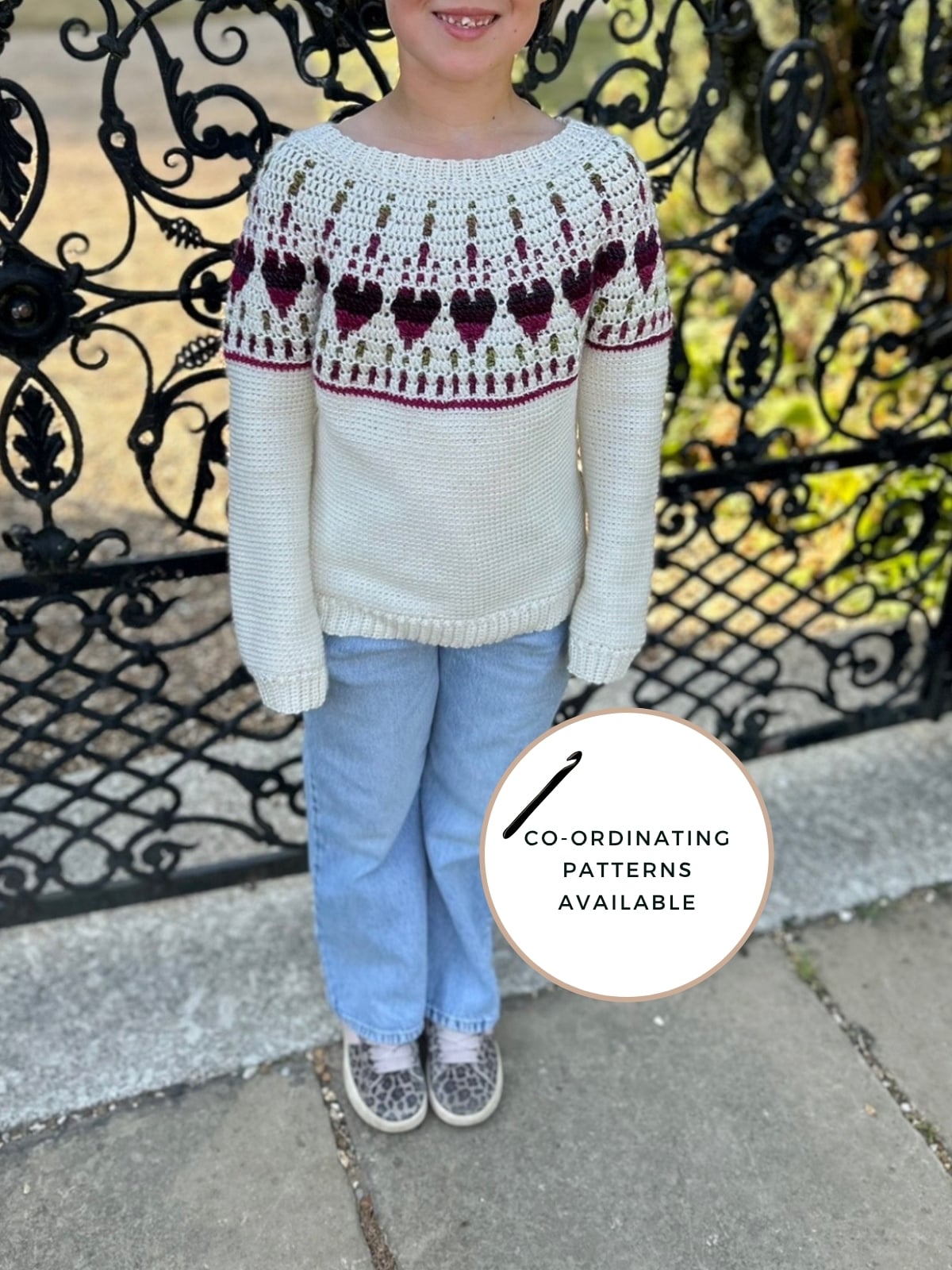 Child standing outside wearing a white crochet sweater with maroon heart pattern, reminiscent of a charming baby crochet sweater pattern, paired with blue jeans and grey shoes. Ornate black metal gate in background. Circular sign reads “Co-ordinating patterns available.”.
