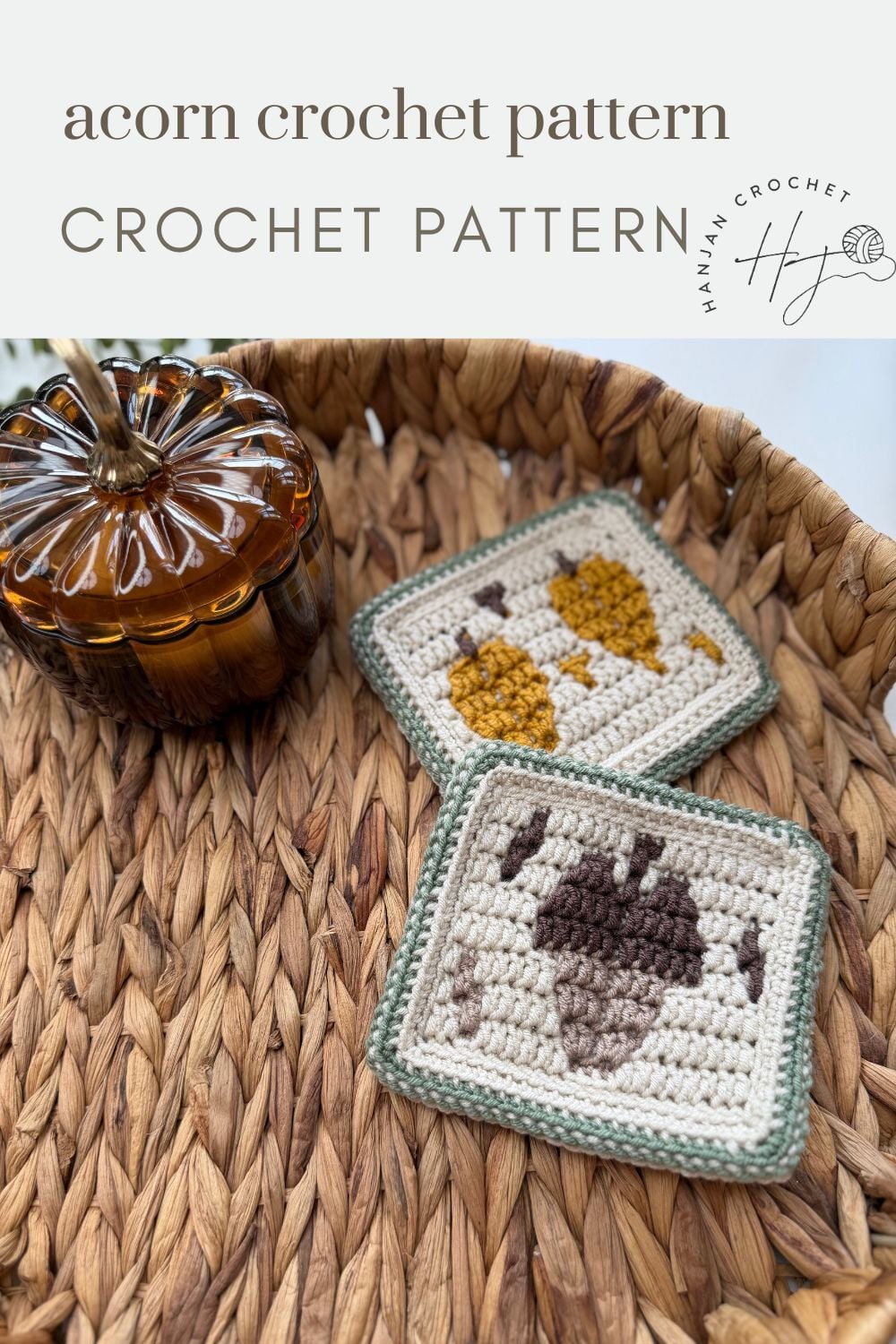Two square crochet coasters, crafted with an intricate acorn crochet pattern and delicate leaf motifs, sit on a woven basket tray beside a small glass pumpkin.