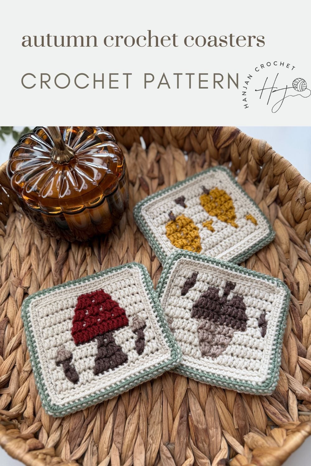 Three square crochet coasters with autumn designs (mushroom, leaves, acorn) are displayed in a woven basket next to a brown glass pumpkin.