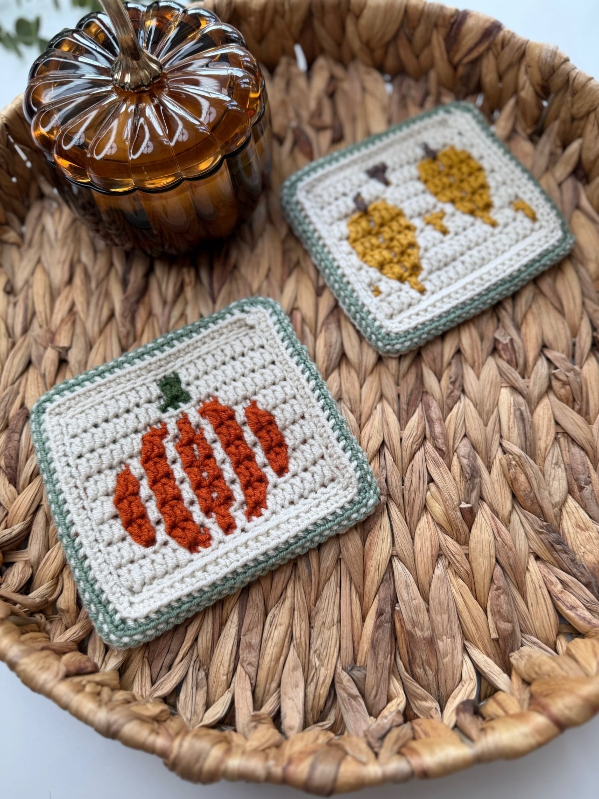 Two crocheted coasters with pumpkin and gourd designs placed in a woven basket next to a glass pumpkin-shaped container.