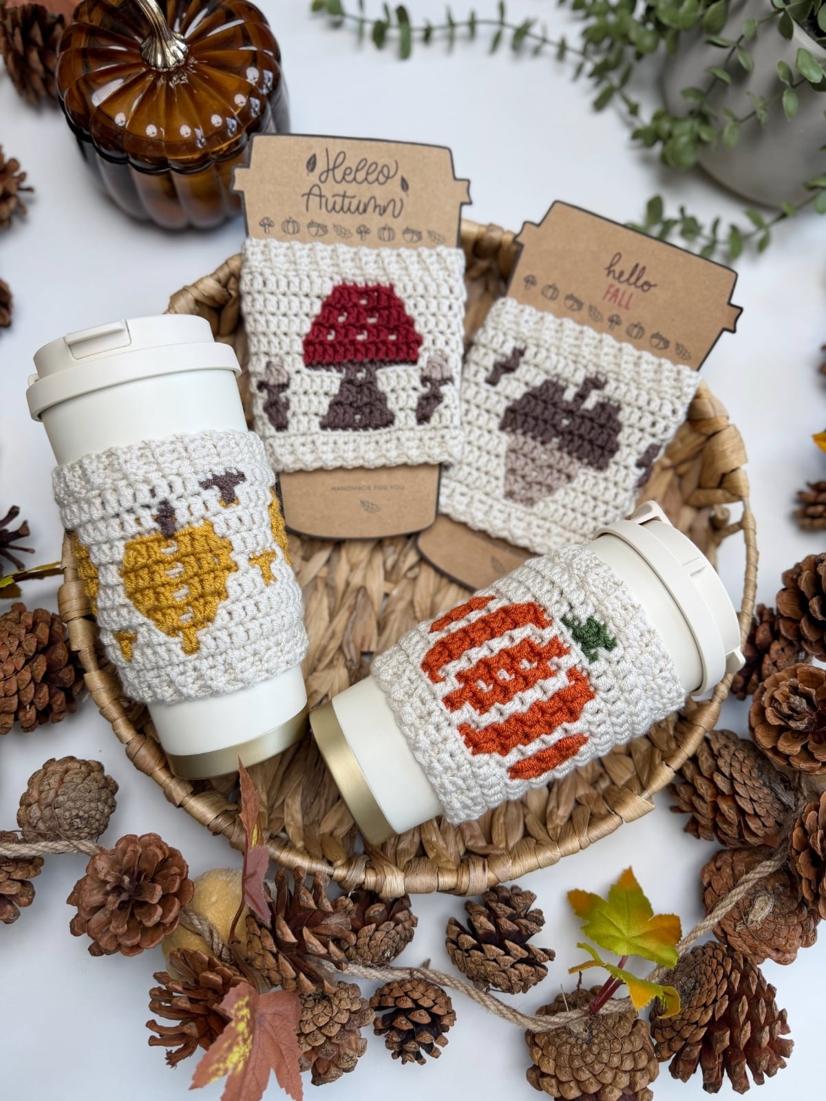 A woven basket with pine cones holds two cups adorned with crocheted autumn-themed cup sleeves—crafted using a charming crochet cup holder pattern—and three matching crochet patches on cardboard, all surrounded by fall leaves and decor.