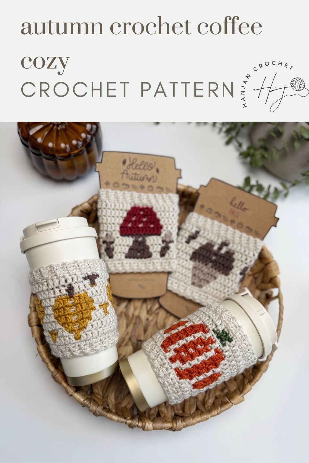 Three reusable cups in crochet cozies with autumn motifs—heart, mushroom, pumpkin—are displayed in a basket, with matching pattern cards behind them, showcasing an adorable crochet cup holder pattern perfect for seasonal sipping.