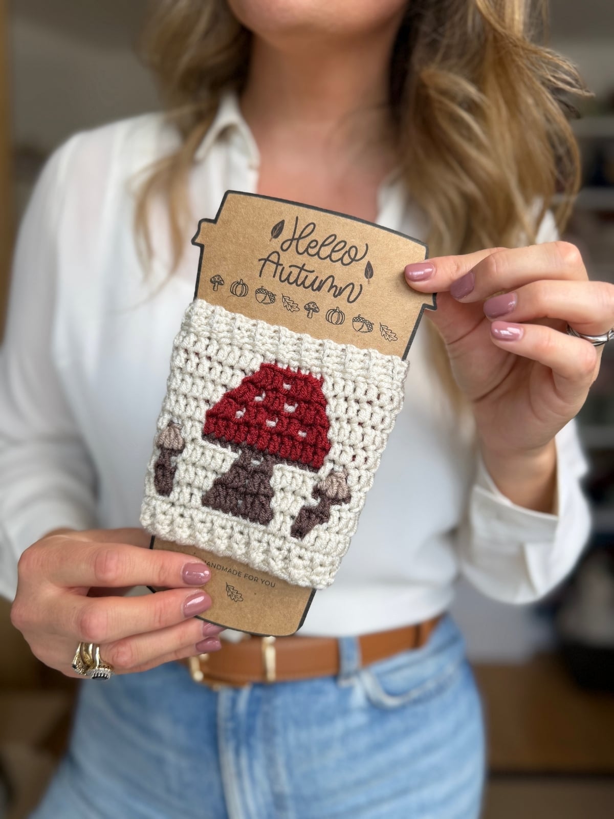 A person holds a crochet cup holder pattern sleeve featuring a crocheted red and brown mushroom design, attached to a cardboard backing labeled “Hello Autumn.”.