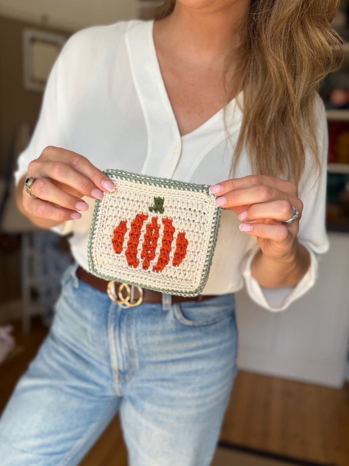 A person in a white top and jeans holds a small crocheted pouch with an orange pumpkin design on the front.