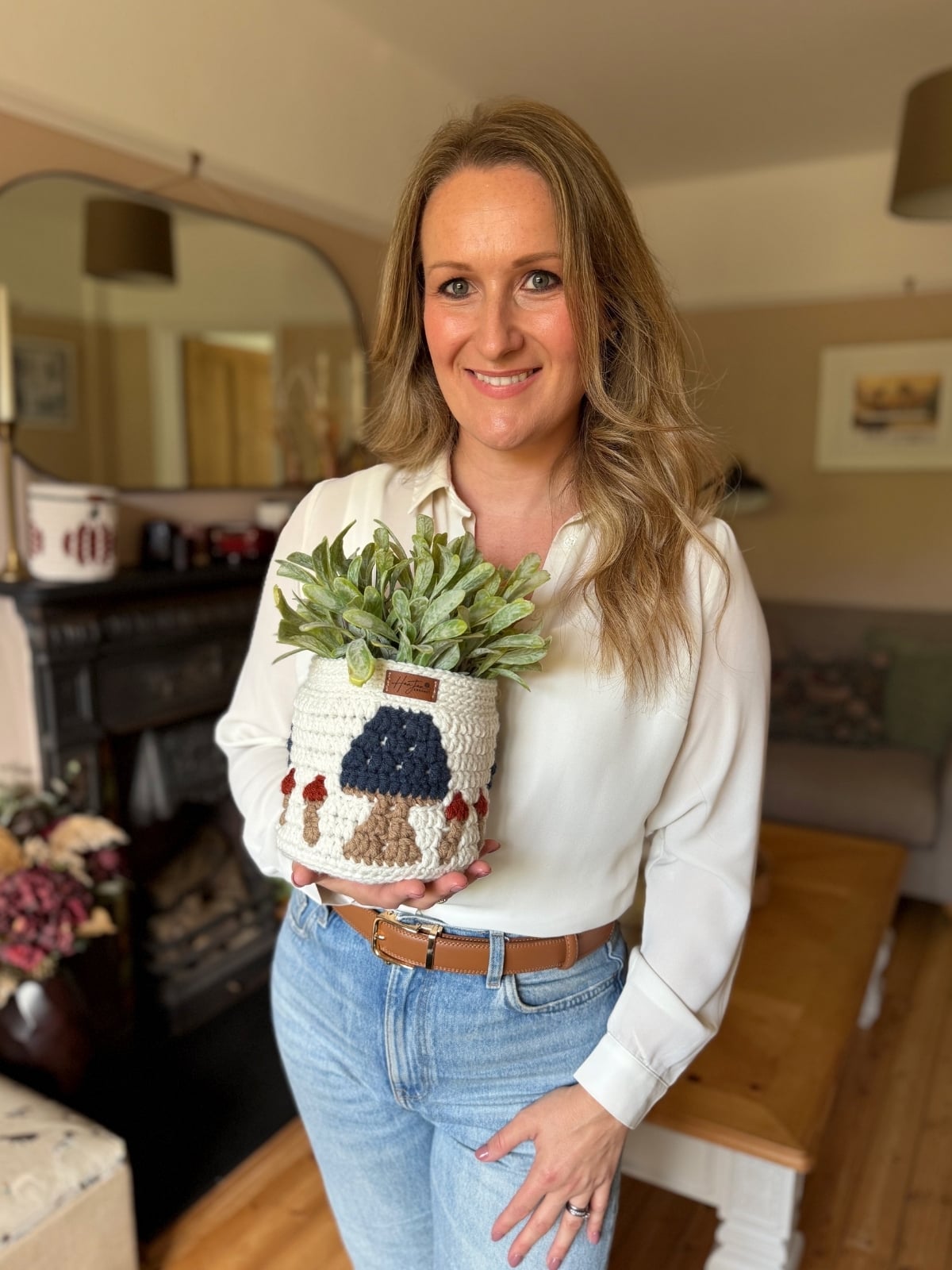 A woman in a white blouse and blue jeans holds a crocheted plant pot with green foliage, standing in a cozy, well-decorated living room that hints at her love for handmade decor and unique crochet storage basket patterns.