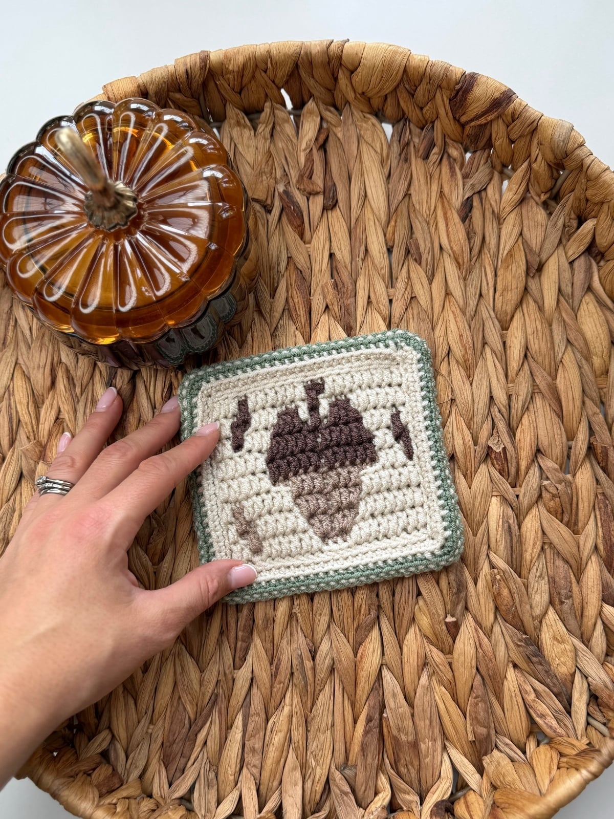 A hand holds a crocheted square featuring an acorn crochet pattern, placed in a woven basket next to a glass pumpkin jar.