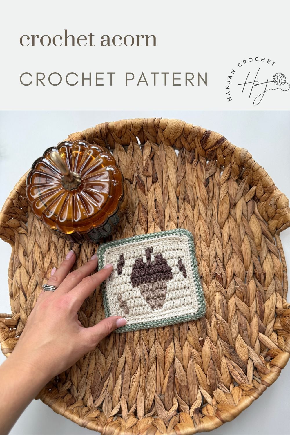 A hand holds a crocheted square featuring an acorn design on a woven tray next to a glass pumpkin; text reads “acorn crochet pattern CROCHET PATTERN.”.