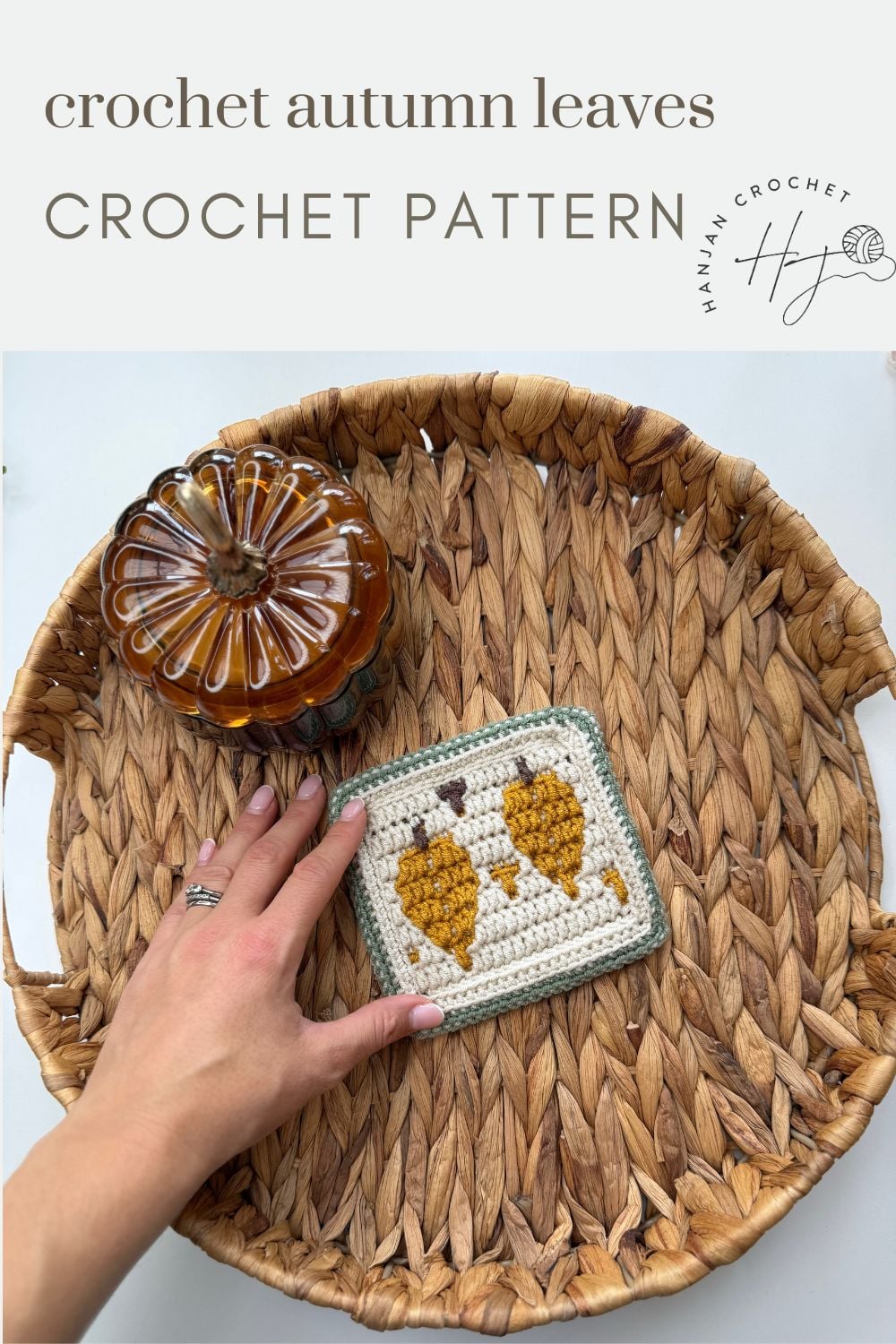 A hand places a crocheted square with two yellow autumn leaves on a woven tray beside a glass pumpkin. Text above reads “crochet autumn leaves CROCHET PATTERN.”.