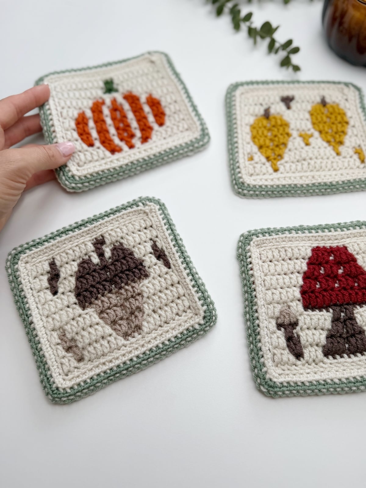 Four crocheted coasters with autumn-themed designs: a pumpkin, two leaves, an acorn, and a red mushroom with white spots. A hand holds the pumpkin coaster. The acorn crochet pattern adds a charming woodland touch to the set.