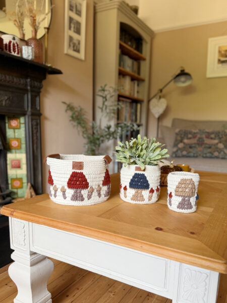 Three crocheted baskets with charming mushroom designs, crafted using a crochet storage basket pattern, sit on a wooden coffee table in a cozy living room with bookshelves and decor in the background.
