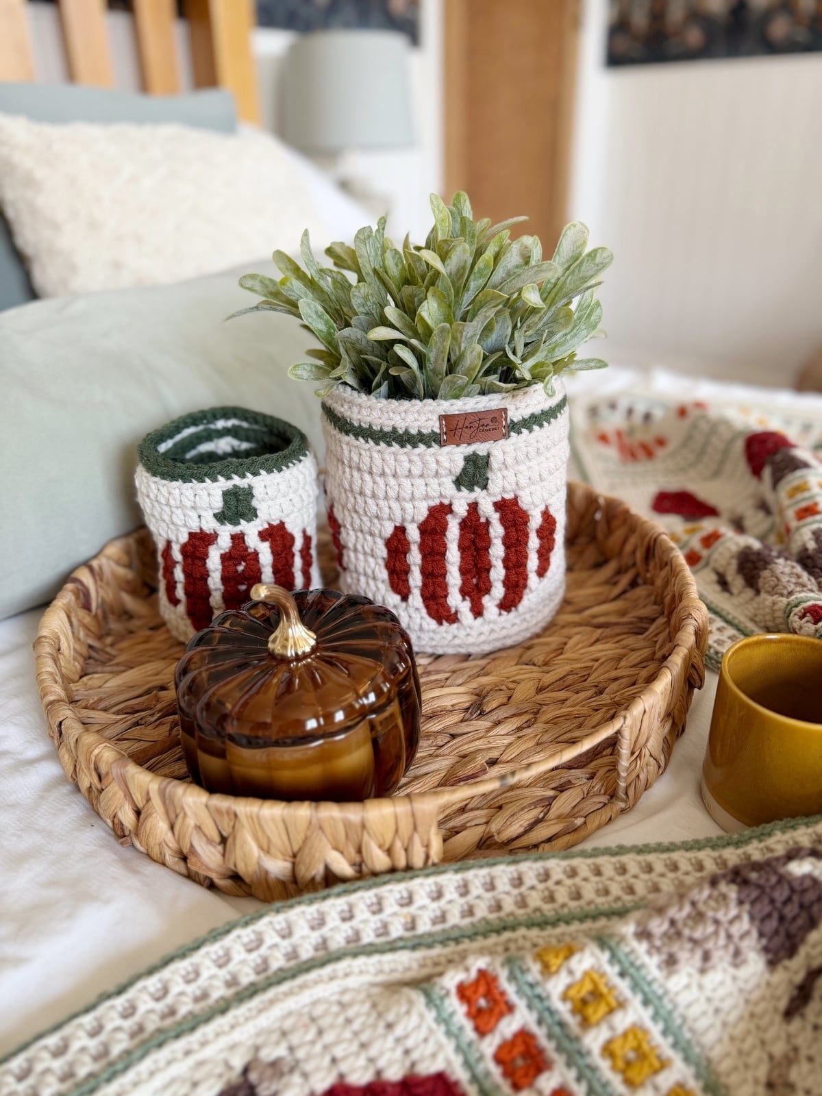 Woven tray with a glass pumpkin jar, a mug, and two crocheted containers—including a charming crochet pumpkin basket holding a potted plant—featuring pumpkin patterns, set on a bed with a crocheted blanket.