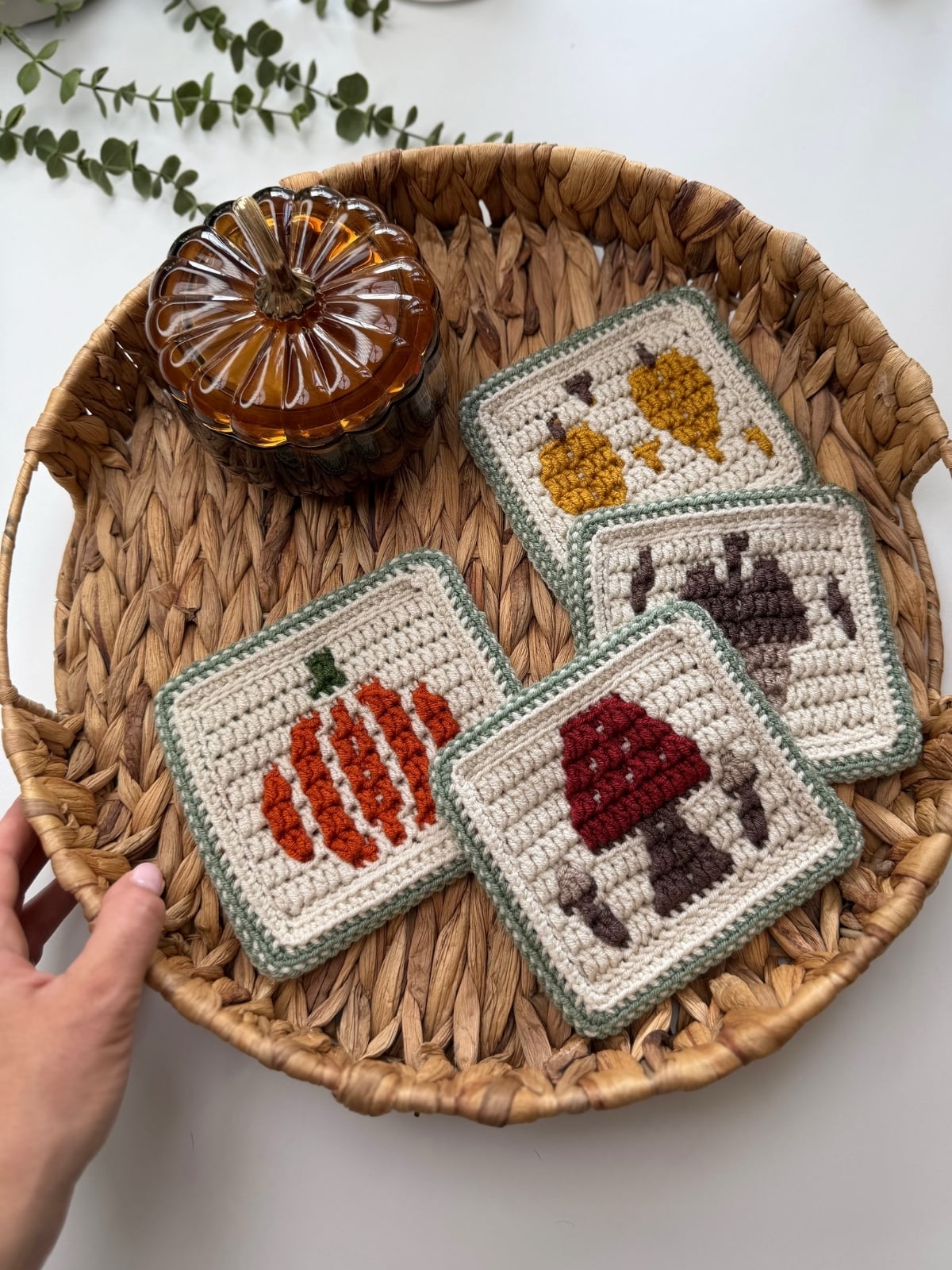 Woven basket with four crochet coasters featuring autumn-themed designs and a small amber glass pumpkin jar. A hand is touching the edge of the basket.