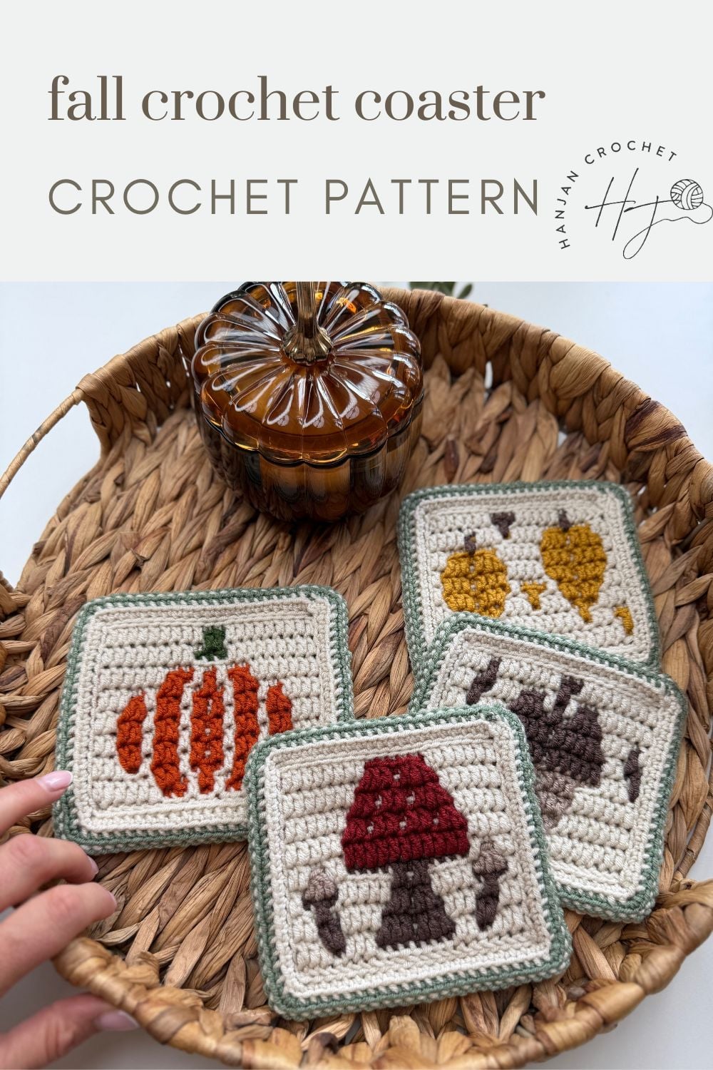 A woven basket holds four crocheted coasters with autumn-themed designs: a pumpkin, leaves, a mushroom, and acorns. A hand reaches for one, and a glass pumpkin jar is in the background.