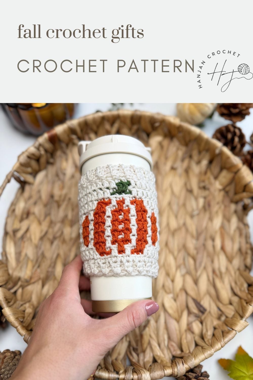Hand holding a cup with a crocheted pumpkin-themed cozy—crafted using a crochet cup holder pattern—placed over a woven tray with pinecones and autumn decor visible around the edges.