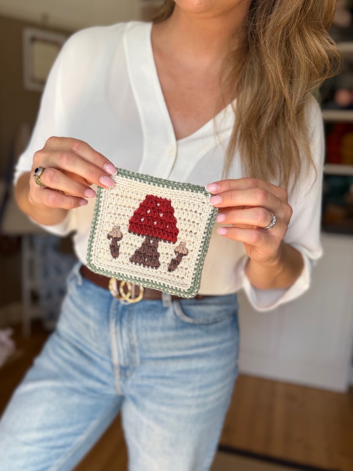 A woman in a white blouse and jeans holds a crocheted square featuring a red and brown mushroom crochet pattern with a green border.