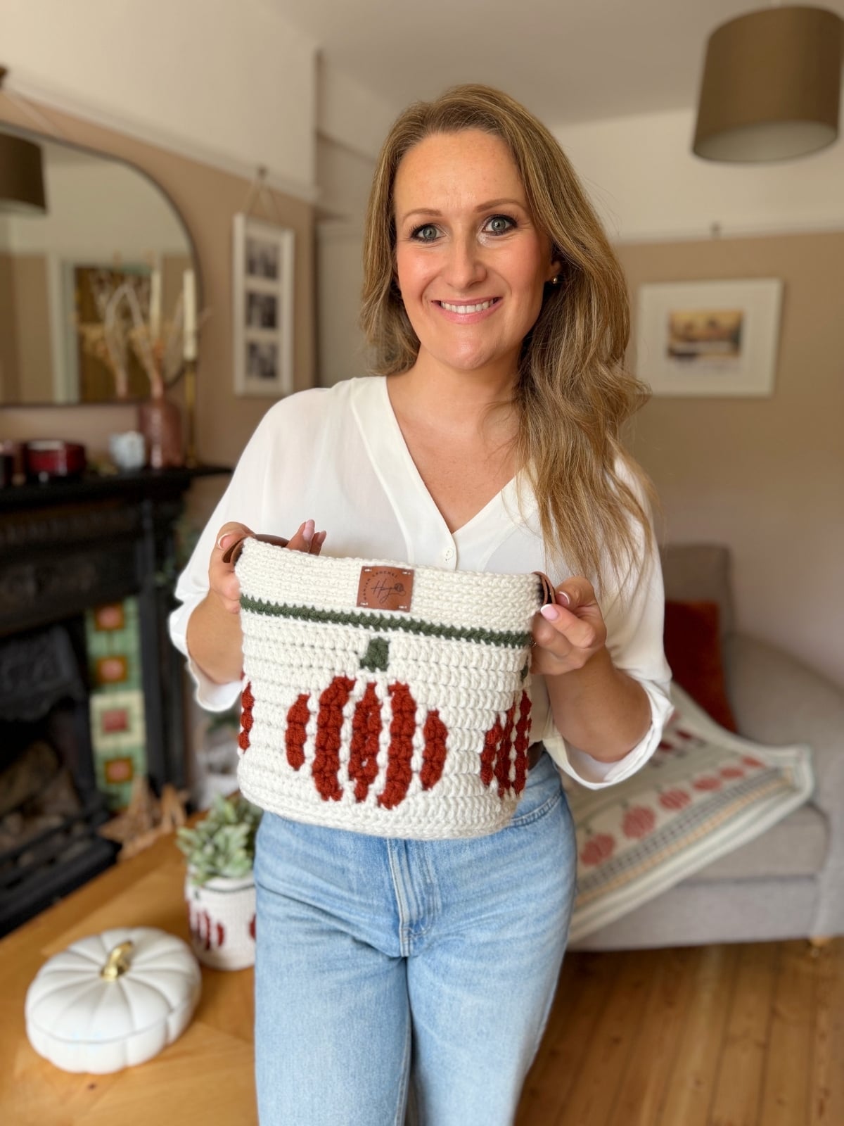 A woman in a white blouse and jeans holds a crochet pumpkin basket in a cozy living room decorated with autumnal accents.